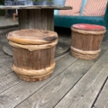 Des paniers en bois transformés en tabourets sur une terrasse en bois, avec une table d'extérieur et un coussin rayé en arrière-plan, évoquent le charme d'un Fût à Saké Japonais des années 1960. Brocante 93