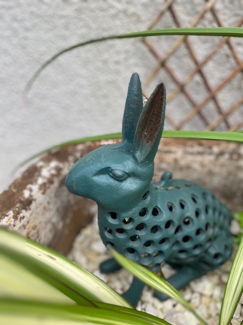 Le Photophore Lapin, une sculpture en métal bleu sarcelle dotée d'un corps perforé, est nichée parmi les plantes vertes du jardin, servant à la fois de pièce décorative enchanteresse et de photophore charmant lorsqu'il est illuminé. Brocante 93