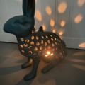 Le Photophore Lapin, lorsqu'il est éclairé de l'intérieur, projette de délicats motifs lumineux à travers ses perforations sur le sol sombre et le fond de la porte blanche. Brocante 93