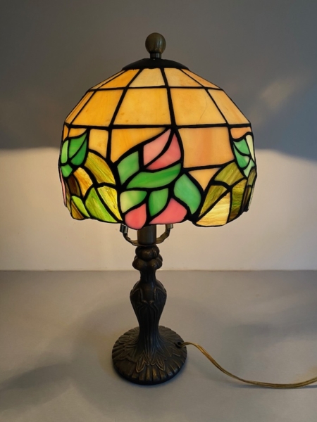 La lampe Tiffany avec vitrail présente des motifs floraux dans des tons de rose, de vert et de jaune sur une élégante base sombre. Brocante 93