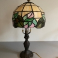 La Lampe Tiffany présente un abat-jour en vitrail orné de superbes motifs floraux, élégamment placé sur une base décorative sombre. Brocante 93