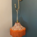 Un abat-jour de style Baladeuse Graphique, orné de couleurs vives et de franges orange, est suspendu élégamment à une applique murale en laiton contre un mur vert foncé. Brocante 93