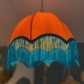 La Suspension Dome Velours, une superbe lampe suspendue orange ornée d'une finition velours et d'une frange bleue, est gracieusement suspendue au plafond, ajoutant du charme à la pièce. Brocante 93