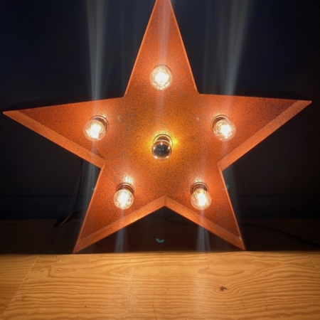 Une Lampe Étoile, un luminaire rustique en forme d'étoile avec des ampoules exposées, projette une lueur chaleureuse sur un fond sombre sur une surface en bois. Brocante 93