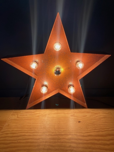 Une Lampe Étoile, un luminaire rustique en forme d'étoile avec des ampoules exposées, projette une lueur chaleureuse sur un fond sombre sur une surface en bois. Brocante 93