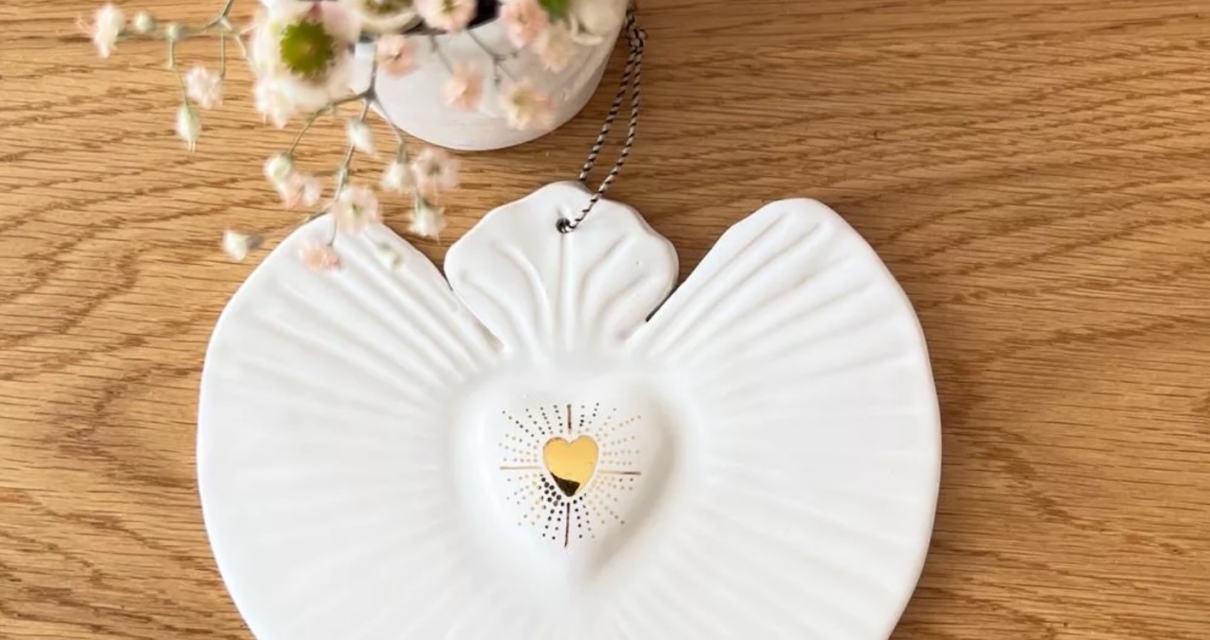 Ornement d'ange en céramique blanche avec un détail de cœur doré, placé à côté d'un petit vase de fleurs sur une surface en bois. Brocante 93