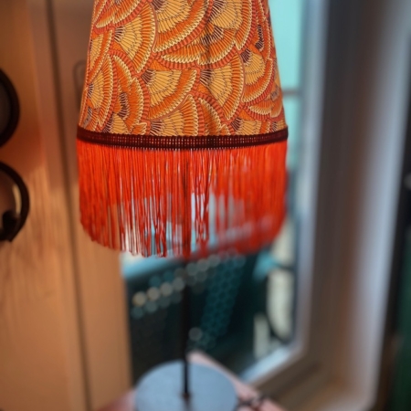 La Lampe Abat-Jour Eventail, dotée d'un abat-jour à motifs orange et jaune vif avec franges, se dresse gracieusement à côté d'une fenêtre. Brocante 93