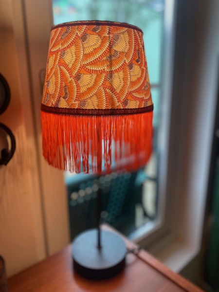 La Lampe Abat-Jour Eventail, dotée d'un abat-jour à motifs orange et jaune vif avec franges, se dresse gracieusement à côté d'une fenêtre. Brocante 93