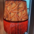 La Lampe Abat-Jour Eventail, avec son motif de plumes orange vif et sa frange rouge ludique, orne une fenêtre, projetant une lueur chaleureuse. Brocante 93