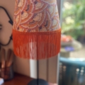 Une Lampe Abat-Jour Eventail dotée d'un abat-jour et d'une frange à motifs de plumes orange complexes est élégamment placée près d'une fenêtre. Brocante 93