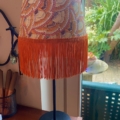 La Lampe Abat-Jour Eventail avec son abat-jour à motifs et sa frange orange se dresse sur une table près d'une fenêtre donnant sur le jardin. Brocante 93