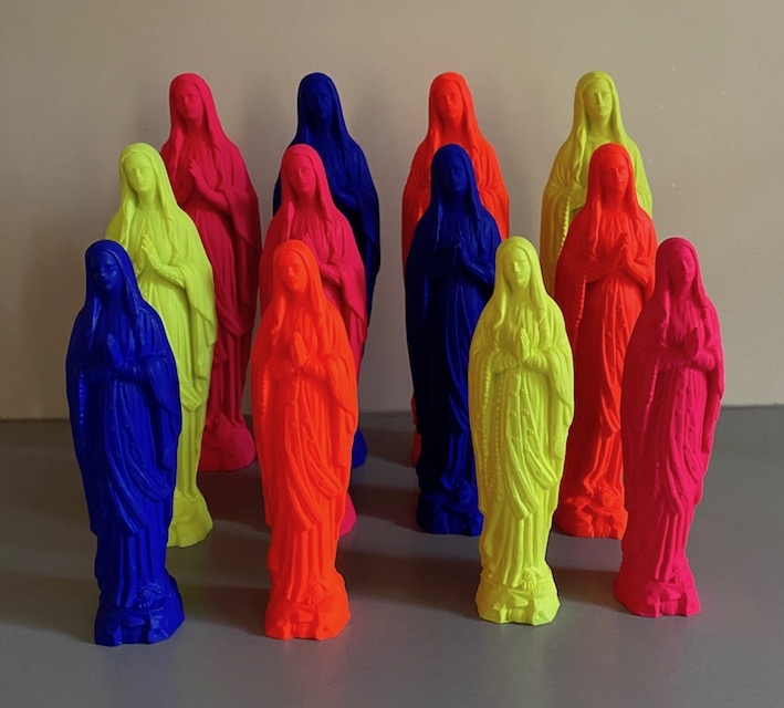 Des statues colorées de personnages en robe dans diverses nuances de néon sont disposées sur une surface plane sur un fond uni. Brocante 93