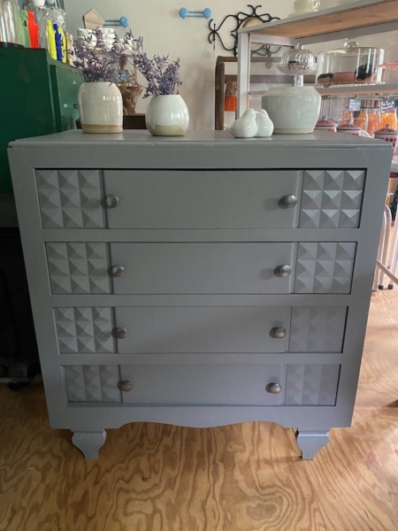 La Commode Grise 1950, aux motifs géométriques sur les tiroirs et surmontée de vases et d'une petite plante, orne une pièce au parquet en bois. Brocante 93