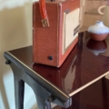 Une radio portable rouge Desserte Vintage 1970 est posée sur une table en bois à côté d'un petit vase avec une fleur violette. Brocante 93