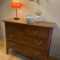 La Commode Américaine 1930, charmante pièce en bois à quatre tiroirs, trône élégamment dans une pièce cosy, décorée d'un abat-jour rouge, de deux vases et de lavande séchée. Brocante 93