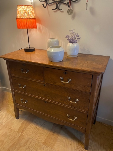 Une commode américaine des années 1930 agrémente la pièce, avec ses tiroirs et sa lampe surmontée d'un abat-jour à franges rouges. Des vases en céramique ajoutent du charme, tandis que des crochets muraux complètent cet ensemble de meubles anciens. Brocante 93