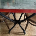 La table Bistrot 1950, dotée d'un plateau en métal, d'un bord rouge vif et de pieds noirs élégants, trône au milieu de chaises en bois sur un plancher en bois, incarnant l'élégance intemporelle du design des années 1950. Brocante 93