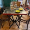 La Table Bistrot 1950 est installée dans un bistrot vintage d'inspiration années 50 avec un plancher en bois, comprenant une table carrée et deux chaises, accentuées par deux bouteilles Ricard vides et des plats jaunes qui ajoutent une touche de couleur. Brocante 93