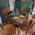 Table de bistrot 1950 avec chaises en bois, trois bouteilles de Ricard et plats en verre colorés dans un cadre cosy. Brocante 93