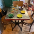 L'ensemble Table Bistrot 1950 présente un design rétro avec des bouteilles en verre et des cendriers colorés, complétés par des chaises en bois dans une pièce vintage classique des années 1950. Brocante 93