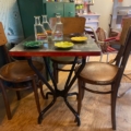 La charmante Table Bistrot 1950 et ses quatre chaises en bois ornent le plancher de bois, orné de plateaux de vaisselle et de bouteilles colorées. Brocante 93