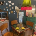 Un espace repas vintage cosy met en valeur la Table Bistrot 1950, ses chaises en bois, sa lampe verte et ses charmantes décorations murales. Brocante 93