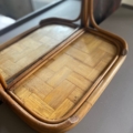 Le Miroir & Etagère Bambou 1960, un petit miroir en bois courbé, est élégamment placé sur une surface à motifs en osier au sommet d'une table sombre. Brocante 93