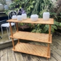 Une Etagère Bambou 1960 présente avec élégance des pots en céramique blanche sur une terrasse en bois, au milieu d'une verdure luxuriante et d'une chaise blanche classique. Brocante 93