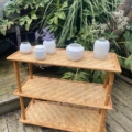 L'Étagère Bambou 1960 évoque une ambiance des années 1960, avec des étagères en osier aux côtés de plantes vertes luxuriantes sur une terrasse en bois, enrichie de pots en céramique blanche et d'accents en bambou. Brocante 93