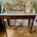 La Coiffeuse double 1940 présente une coiffeuse en bois avec un plateau en marbre, ornée d'élégants présentoirs à bijoux et colliers sur bustes, posée sur un plancher en bois rustique. Brocante 93