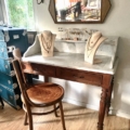 La Coiffeuse double 1940 présente une coiffeuse en bois vintage avec deux présentoirs à bijoux, une chaise élégante et un miroir mural, le tout doucement éclairé par la lumière de la fenêtre venant de gauche. Brocante 93